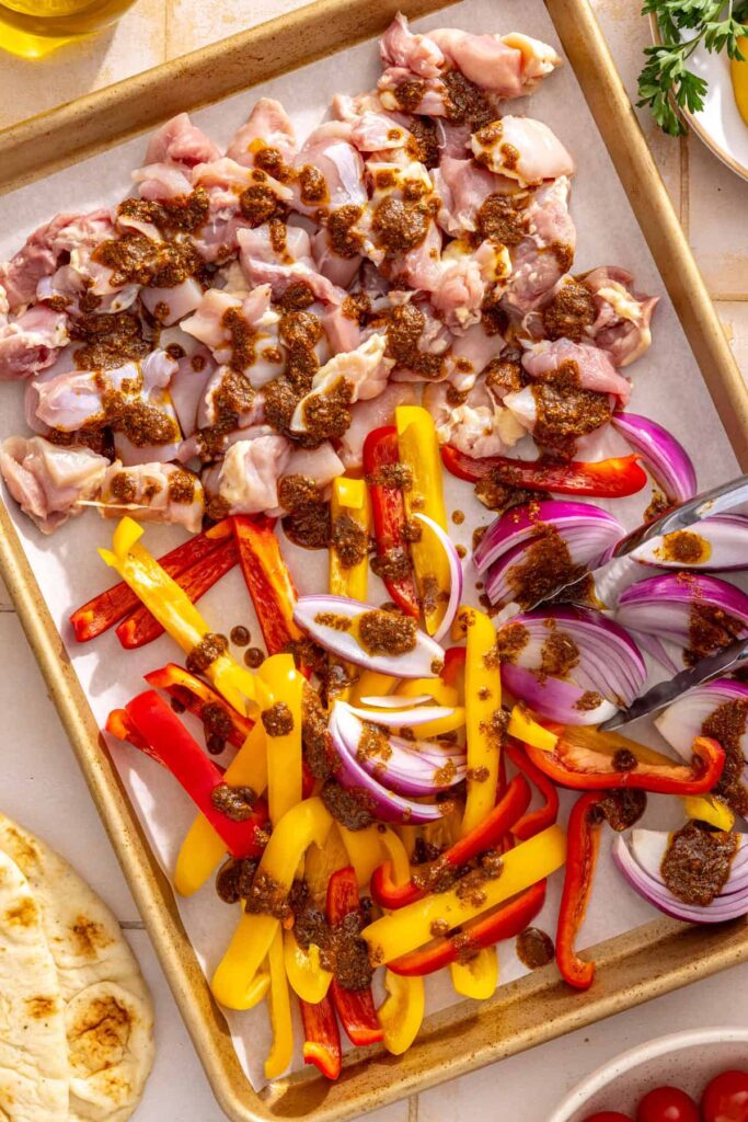 Shawarma marinade poured over raw chicken thighs, bell peppers, and red onion on a sheet pan.