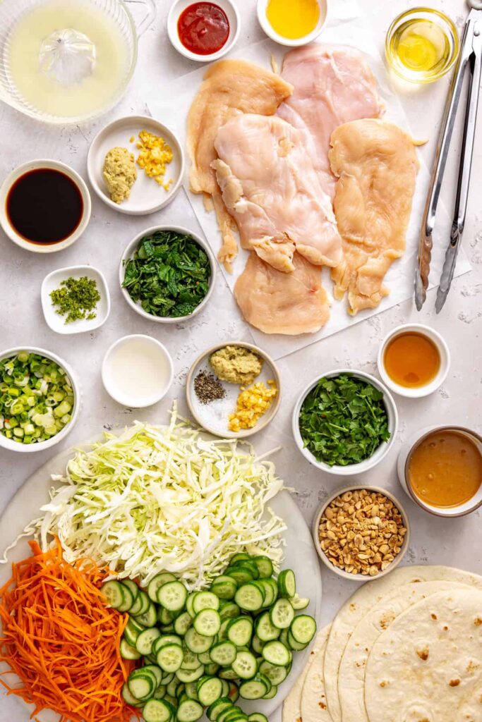 Ingredients for Thai peanut chicken wraps, including chicken breast, cabbage, carrots, cucumbers, fresh herbs, peanut butter, lime, and tortillas.