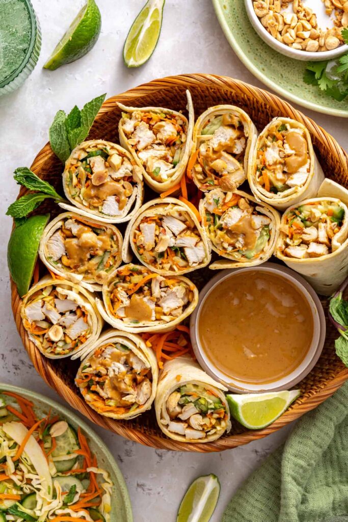 Overhead view of sliced Thai peanut chicken wraps arranged upright on a serving platter with creamy peanut dipping sauce and a lime wedge.