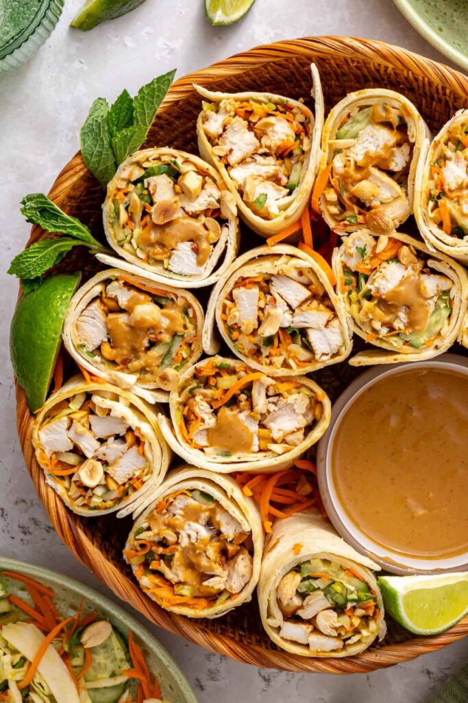 Healthy Thai peanut chicken wraps sliced in half and displayed cut-side up on a platter with fresh greens, lime, and peanut dipping sauce.