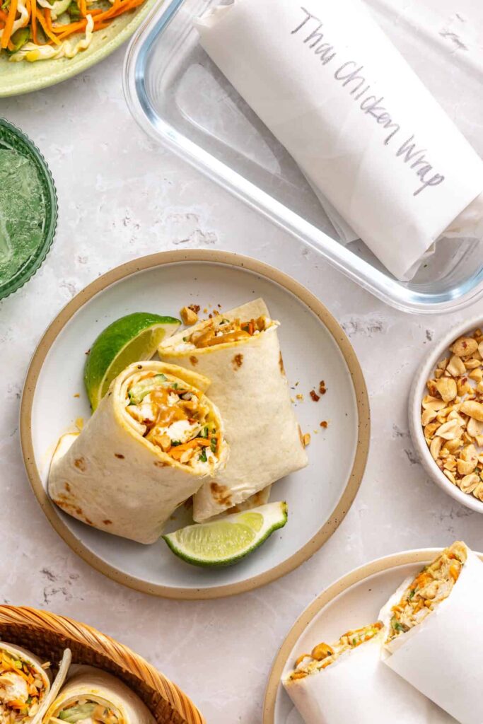Side-angle view of two halves of a Thai peanut chicken wrap stacked on a plate with a lime wedge and drink beside it.