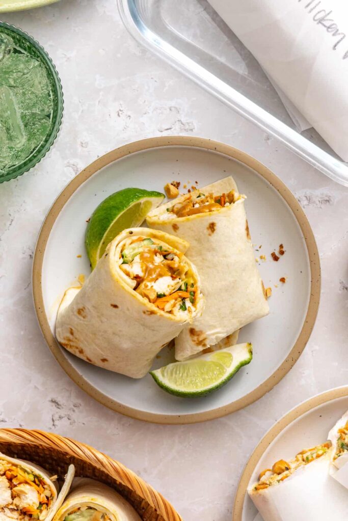Thai peanut chicken wrap sliced into two halves and stacked on a plate with a fresh lime wedge and drink in the background.