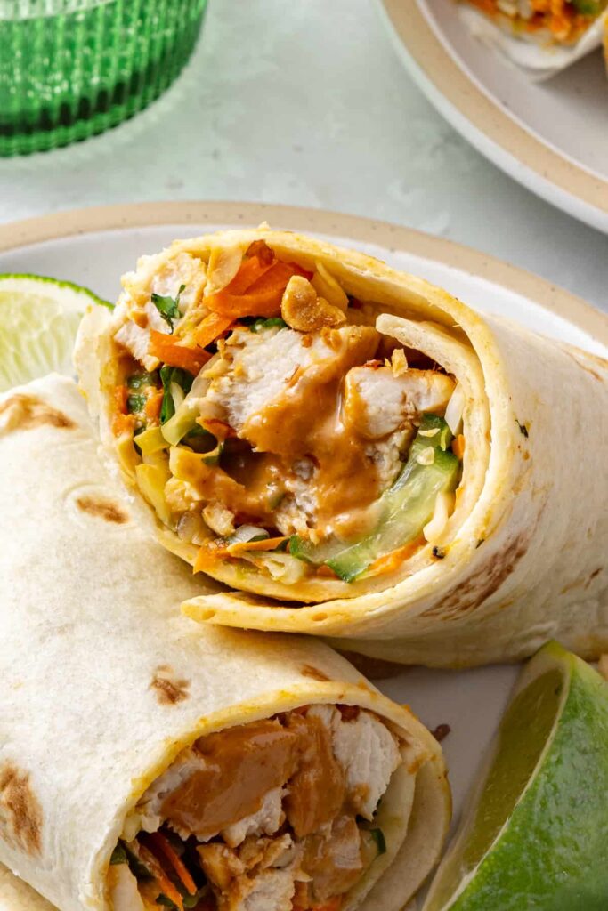 Two stacked halves of a homemade Thai peanut chicken wrap showing the tortilla, marinated chicken, and colorful slaw on a plate.