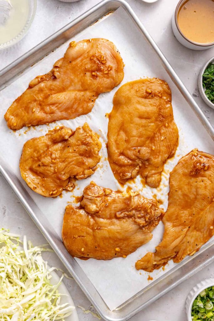 Marinated chicken breasts arranged in a single layer on a parchment-lined baking sheet before baking.